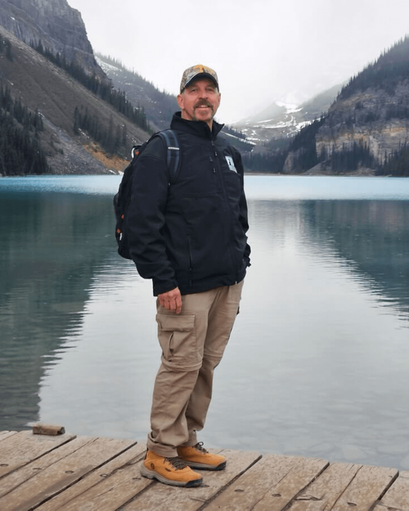 James Roger wearing a backpack and standing on a lake dock with mountains in the background